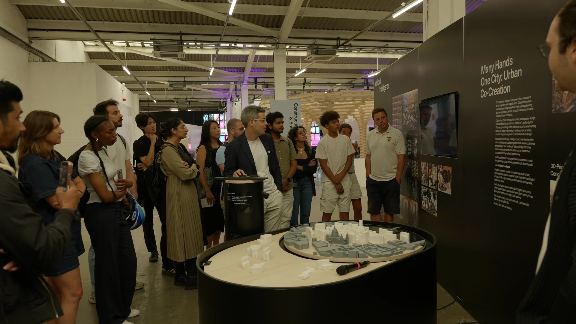 A diverse group of visitors gather around a circular display model in a modern museum or exhibition space with industrial ceiling and informational panels.