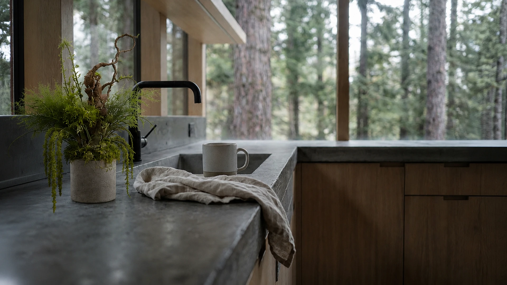 A close-up of a minimalist kitchen featuring a raw concrete countertop, warm natural wood cabinetry, a matte black faucet, handcrafted ceramic tableware, natural linen textiles, sculptural greenery, and expansive glazing that frames a serene forest landscape.