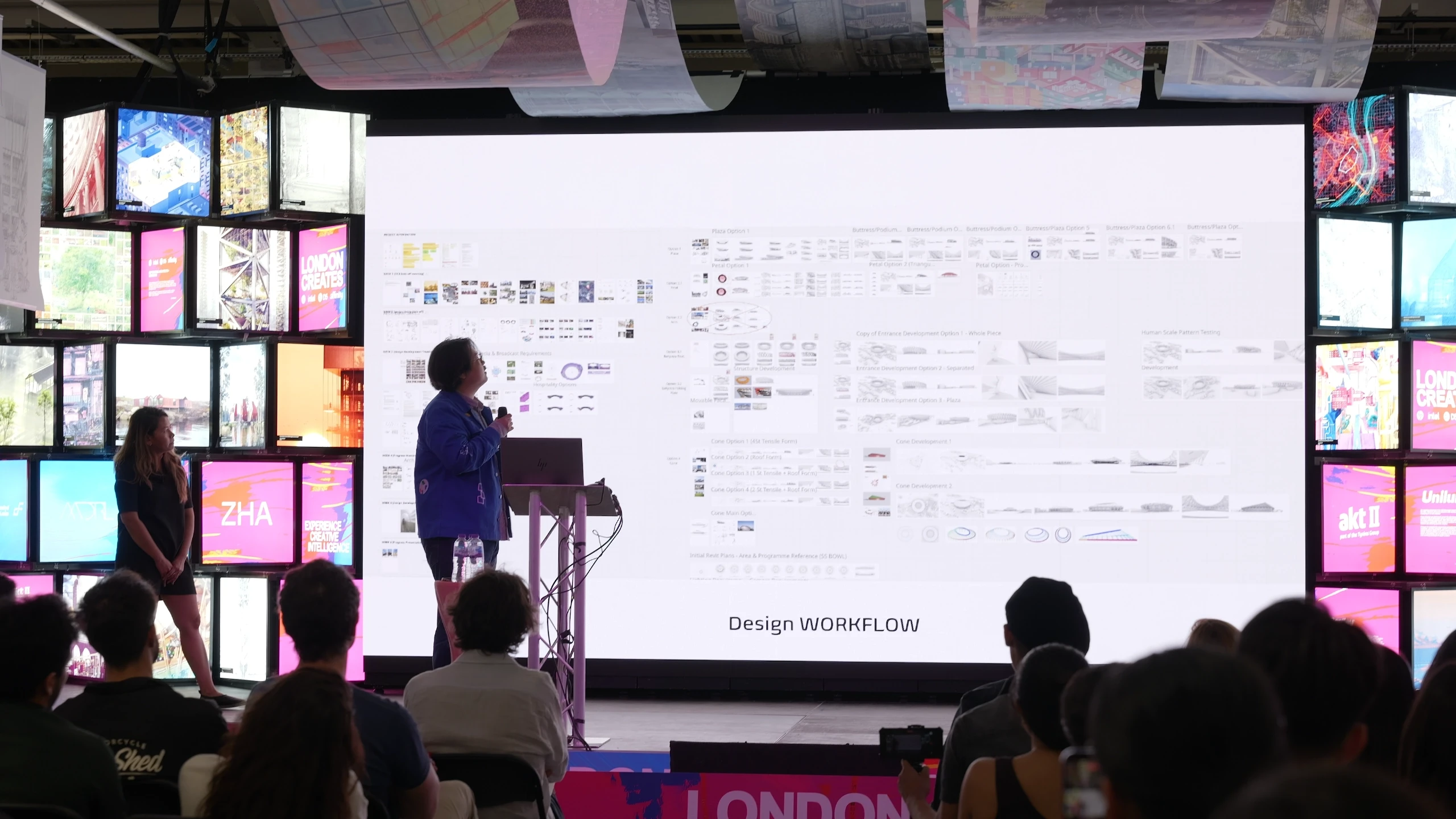 A presenter demonstrates a design workflow on a large projection screen at a conference, with colorful digital displays visible on both sides of the stage.