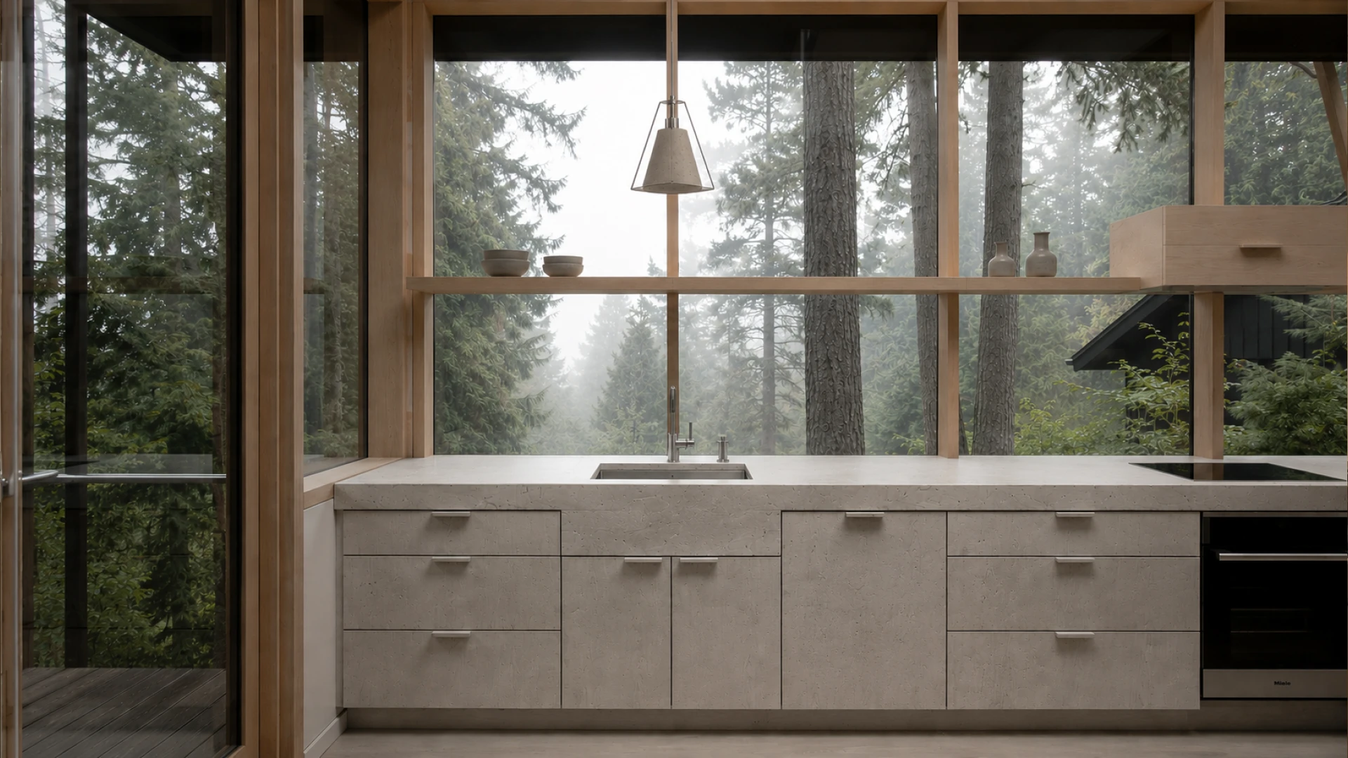 A minimalist kitchen featuring raw concrete cabinetry, a light honed stone countertop, warm natural wood framing and shelving, matte black fixtures, and expansive floor-to-ceiling glazing overlooking a misty forest landscape.