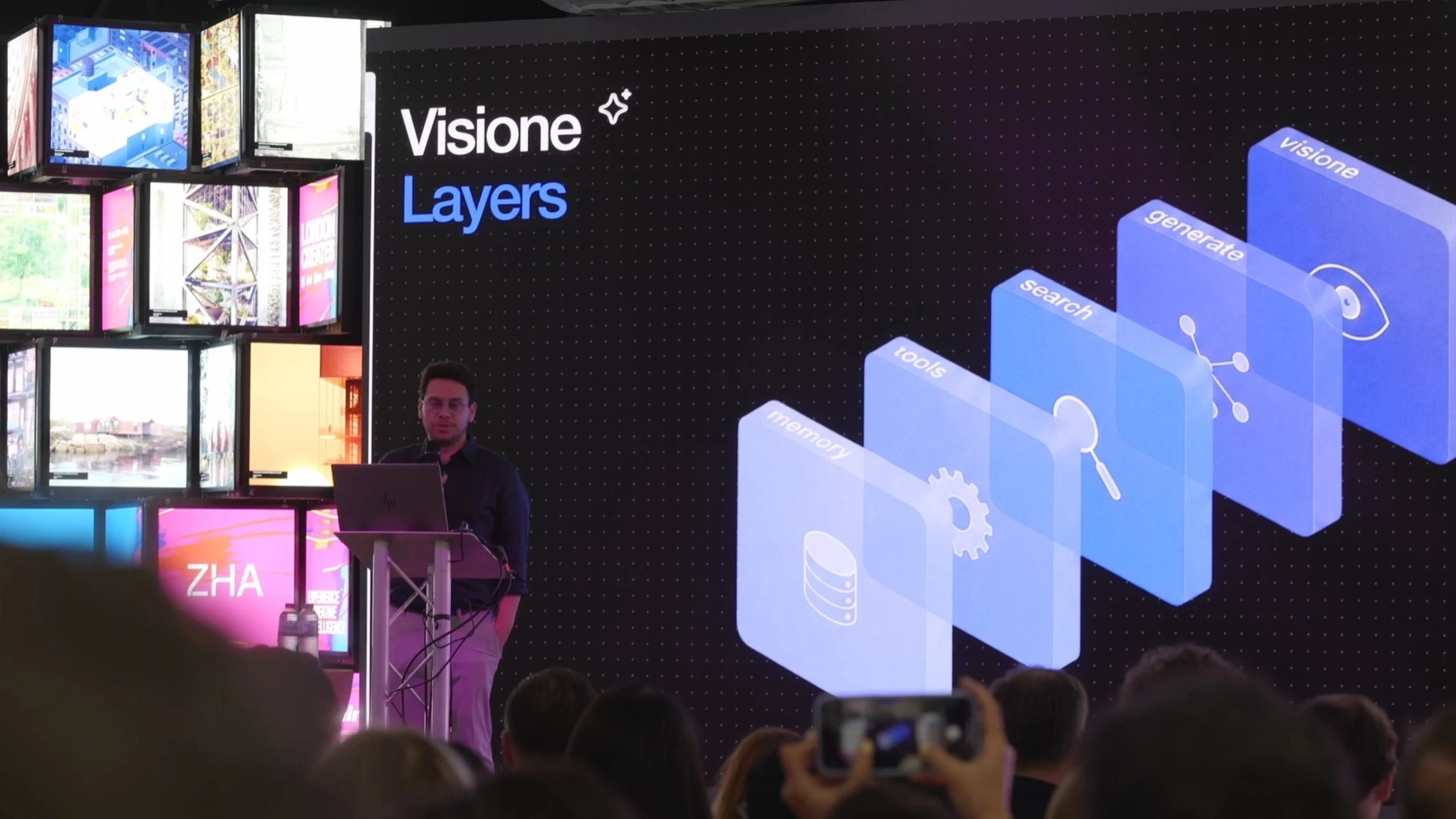 Presenter on stage demonstrating Visione Layers technology with isometric 3D graphics and visual display panels